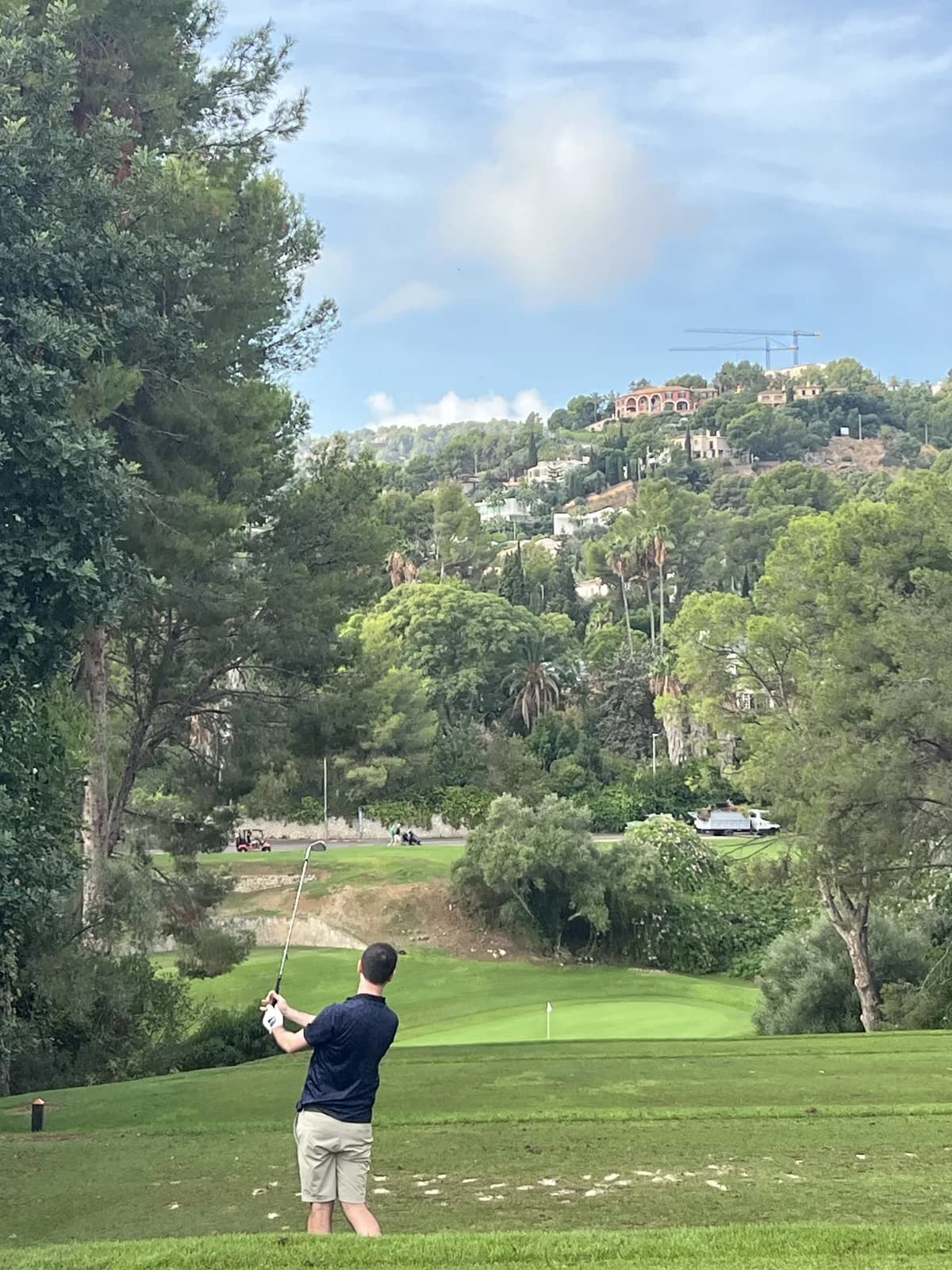 Golfer in a navy polo hitting an iron towards a green with a wooded hillside and houses behind at Son Vida