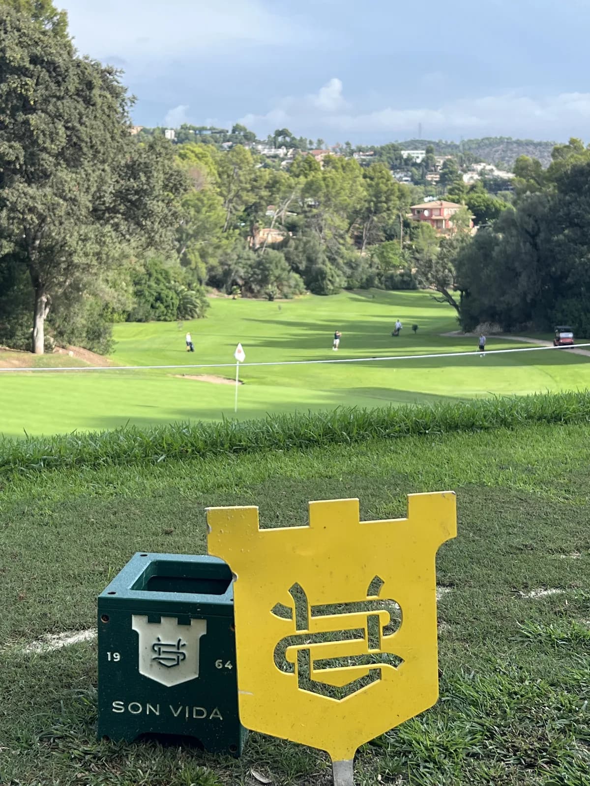 Yellow Son Vida crest tee marker and a green Son Vida 1964 marker on the 12th tee, with the fairway of the 11th stretching back below