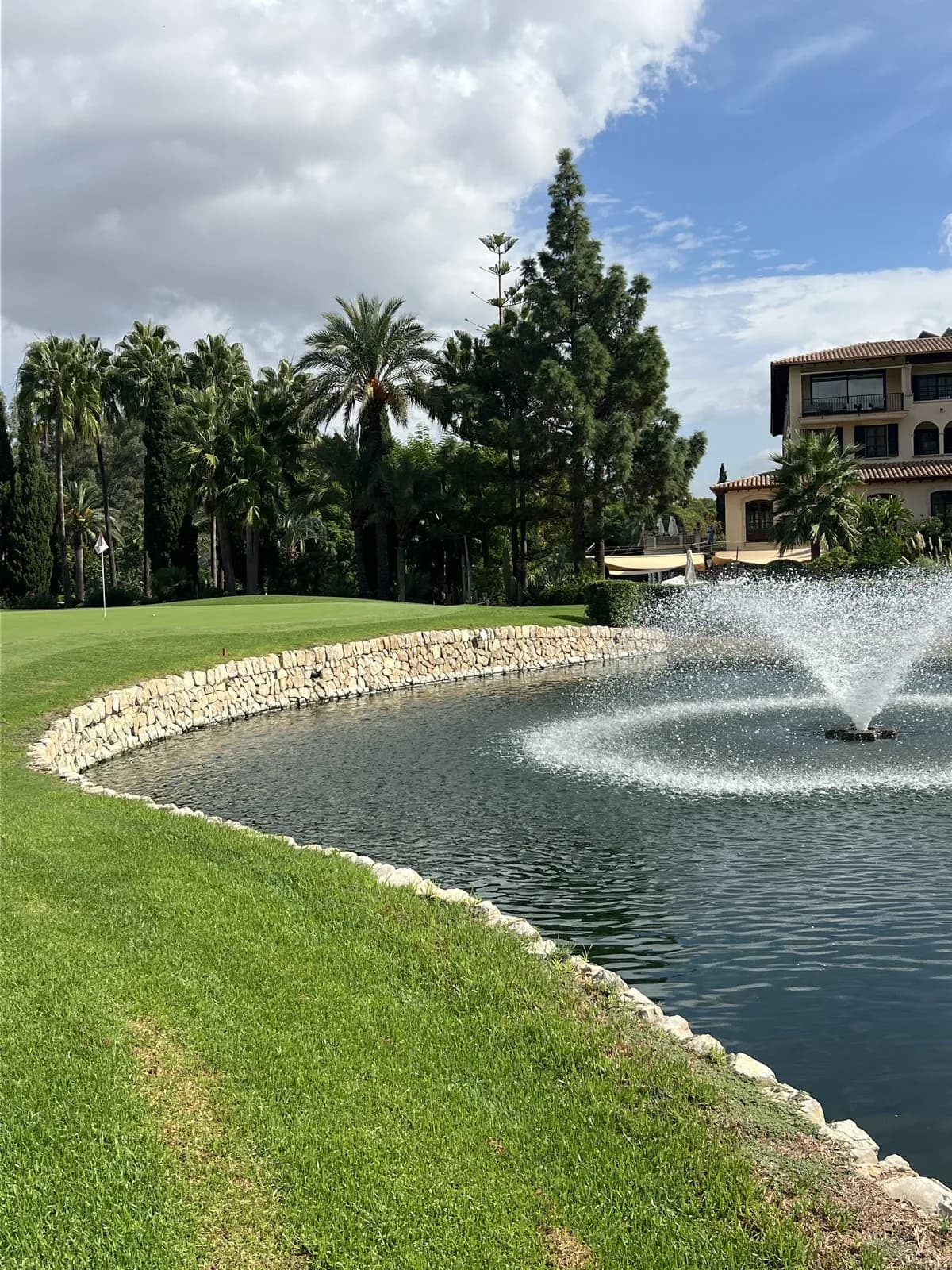 Fountain on a stone-edged lake with the Son Vida hotel behind and palm trees beyond a green
