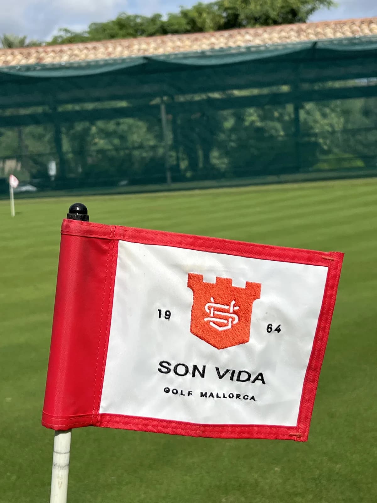Red and white Son Vida 1964 flag on the practice putting green with the practice net behind