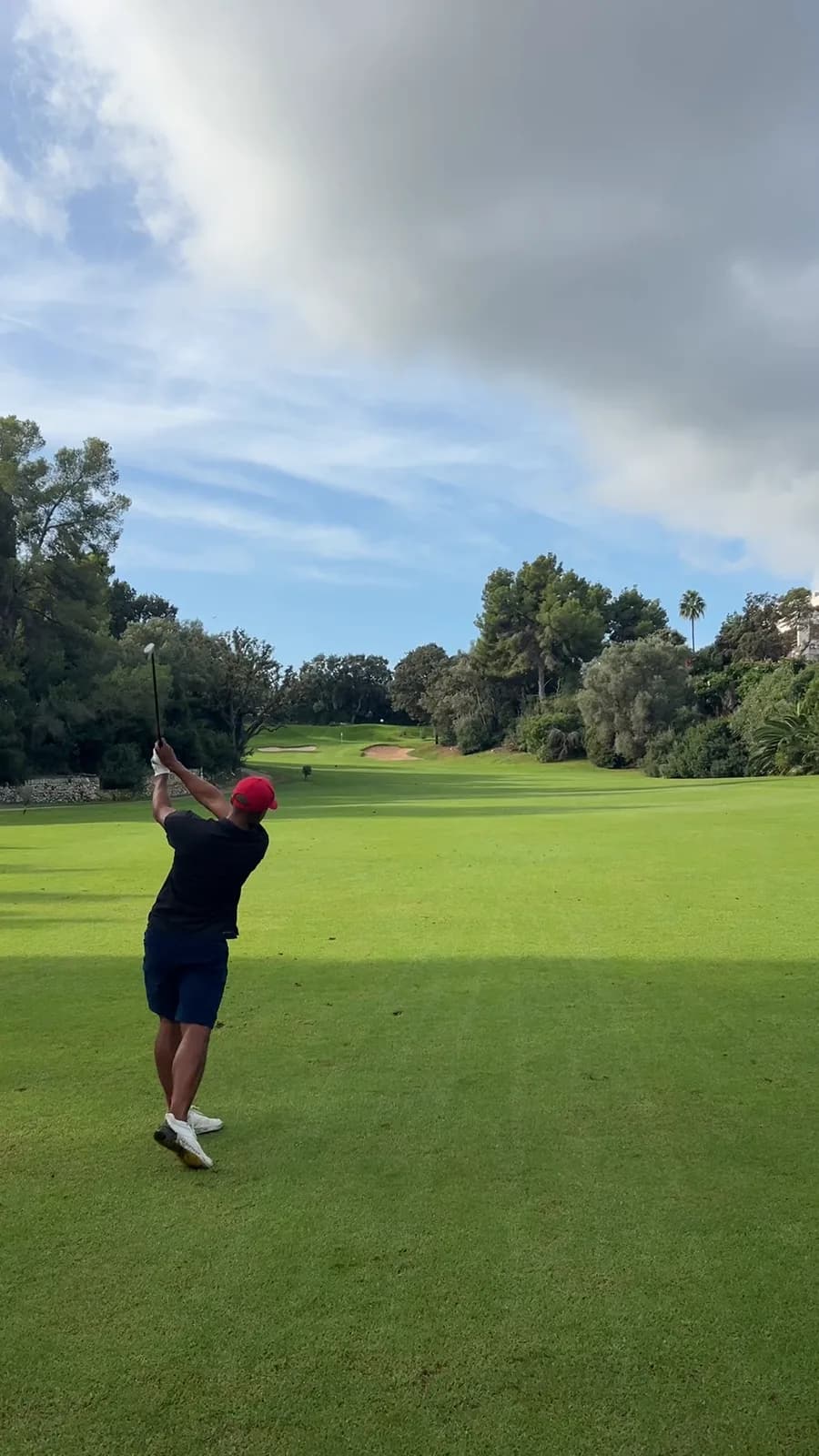 Golfer in a red cap finishing an iron shot from the fairway towards bunkers and the green on the par 5 11th at Son Vida